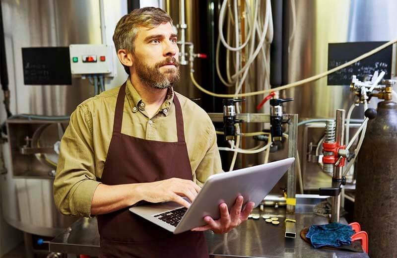 A brewer in the back house of brewery examining tanks and beer making process while using a laptop with Ollie