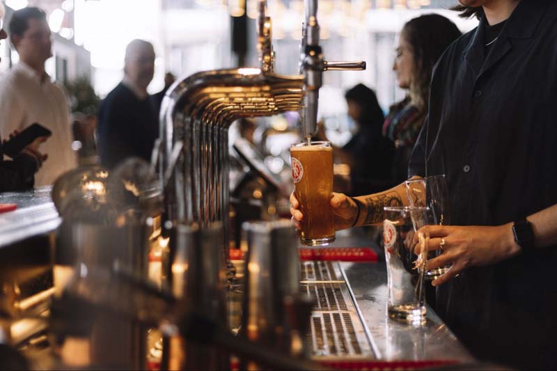 A close up photo of a busy bar and bartender pouring beer into a branded glass from tap