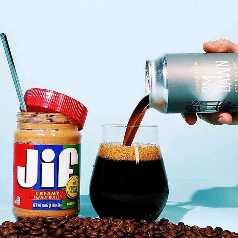 A can of PM Dawn coffee beer poured into a glass with coffee beans and a jar of JIF peanut butter