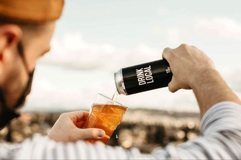 A person pouring a can of craft that reads "Drink Local" into a glass outdoors
