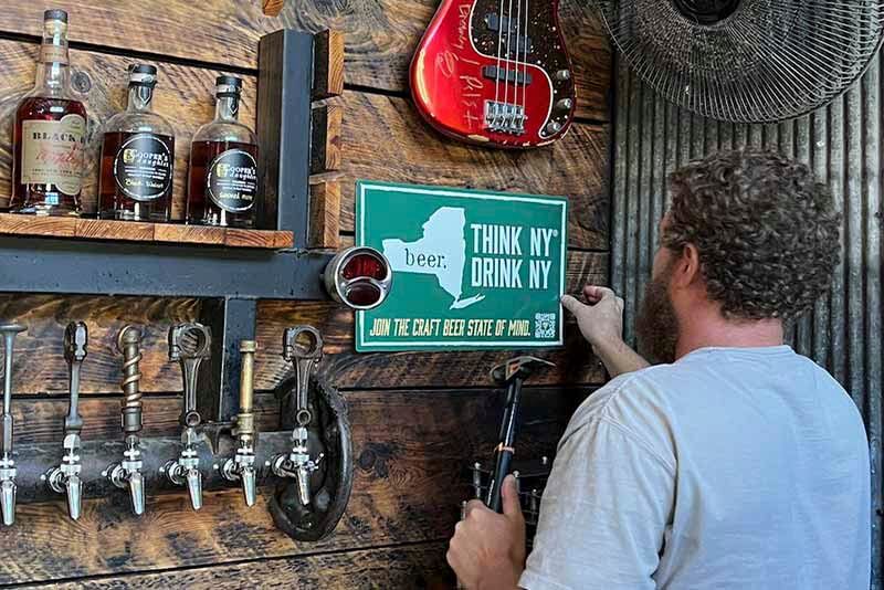 A brewers hammers a sign saying "Think NY, Drink NY" the slogan for the New York State Brewers Association next to a wall of craft beer taps