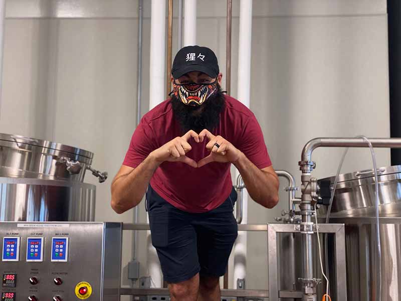 Shojo Beer co brewer making a hand heart symbol in brew house in front of brew kettles